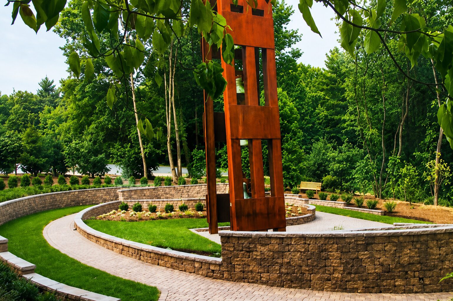 Rust-colored abstract monument rises from a circular landscaped garden, bordered by stone walls and paths, surrounded by lush green trees.